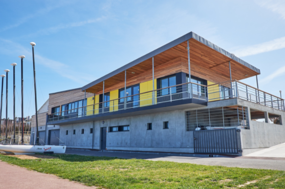 The sailing school building: modern architecture with rough concrete walls for the ground floor, topped by a terrace and a wooden building with a cantilevered roof designed to provide shade for visitors.