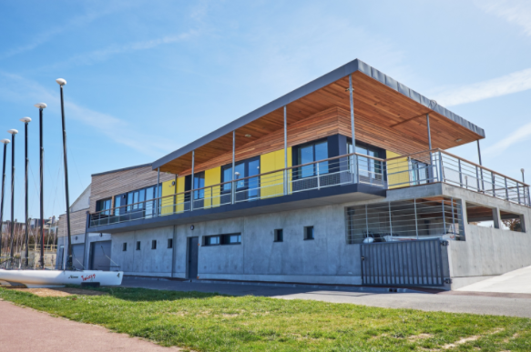 Le bâtiment de l'école de voile : une architecture moderne au murs de bétons brutes pour le rez-de chausé, surmonté d'une terrasse et d'un bâtiment en bois dont le toit en porte-à-faux est conçu pour donner de l'ombre aux visiteurs.