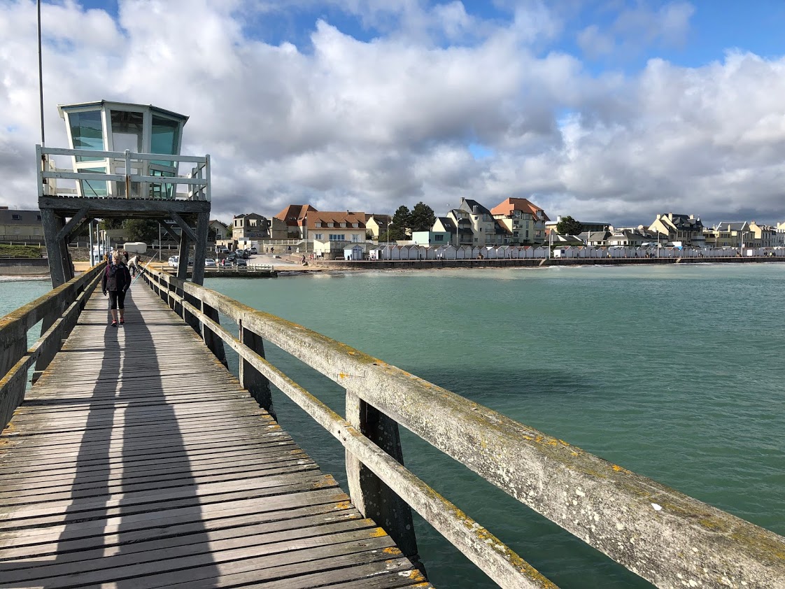 De pier en zijn torentje bij vloed, de blauwe lucht bezaaid met witte en grijze wolken en de stad Luc-sur-Mer in de verte.