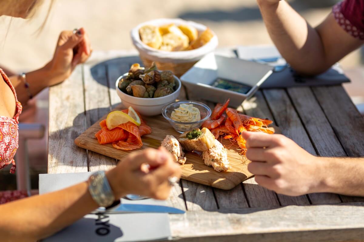 Placed on a wooden table, the Papagayo appetizer board features an assortment of whelks, smoked salmon, shrimp and fish rillettes. The hands of the couple seated at the table are clearly visible.