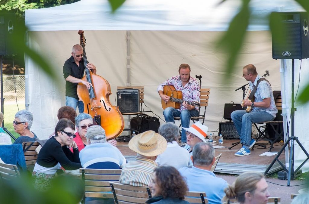 Under a white canvas canopy, a double bass player and two guitarists liven up the Luc-sur-Mer aperitif concert. Sitting on the crates set up for the concert, the audience listen and chat.