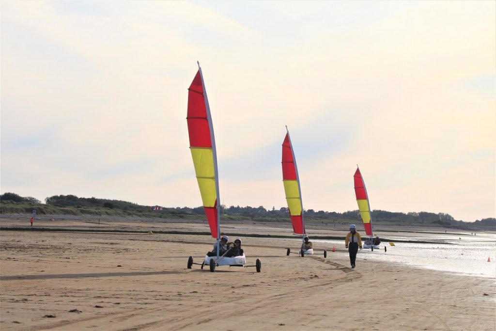 On the beach at Courseulles, in the direction of Grays-sur-Mer, at low tide three sailing boats, one after the other, move towards the photographer, their red sails with wide yellow stripes running vertically across the picture. The light is soft, the sky white with clouds, and in the distance the grassy dunes of this part of the coast can be seen.