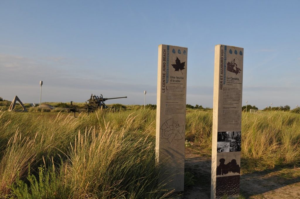 Juno Park, located on the dunes on the left bank of the Seulle. In the foreground, two explanatory panels mark the entrance to the park, and in the background, behind the tall grass and grasses, a tetrahedron (on the left) and an allied cannon, relics of the Normandy Landings.