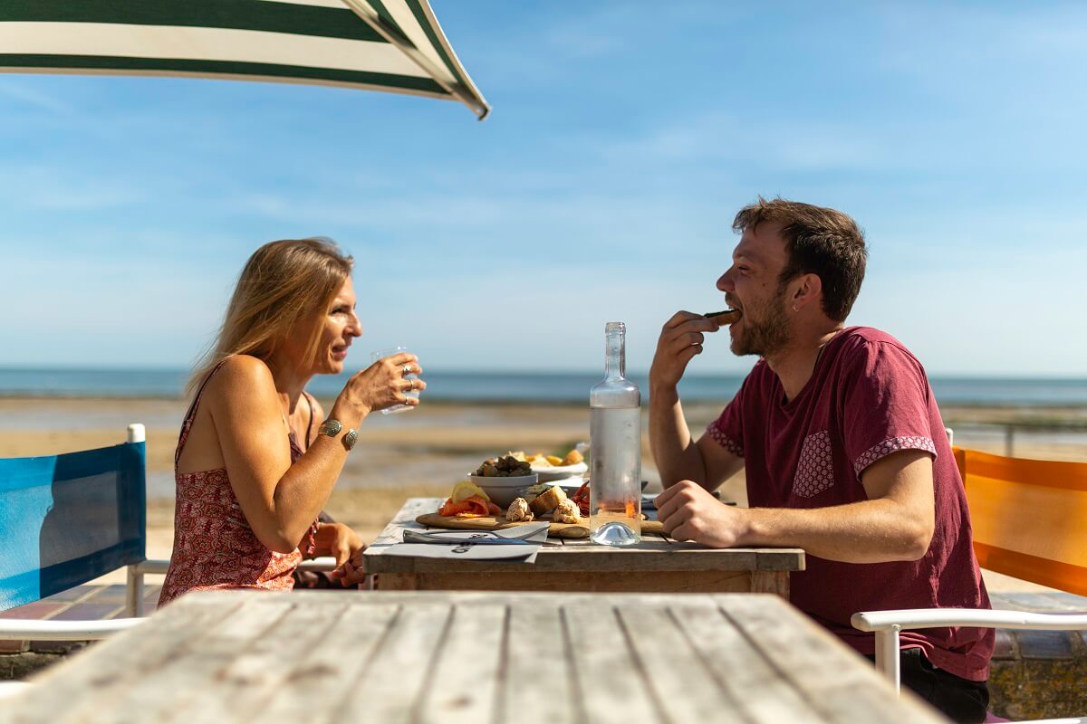 Sur la digue, devant la plage, un jour de beau temps, assis sur l'une sur un siège bleu, l'autre sur un siège orange, abrité par un parasol vert et blanc, un couple déjeune de bon appétit au Papagayo, restaurant de Saint-Aubin-sur-Mer