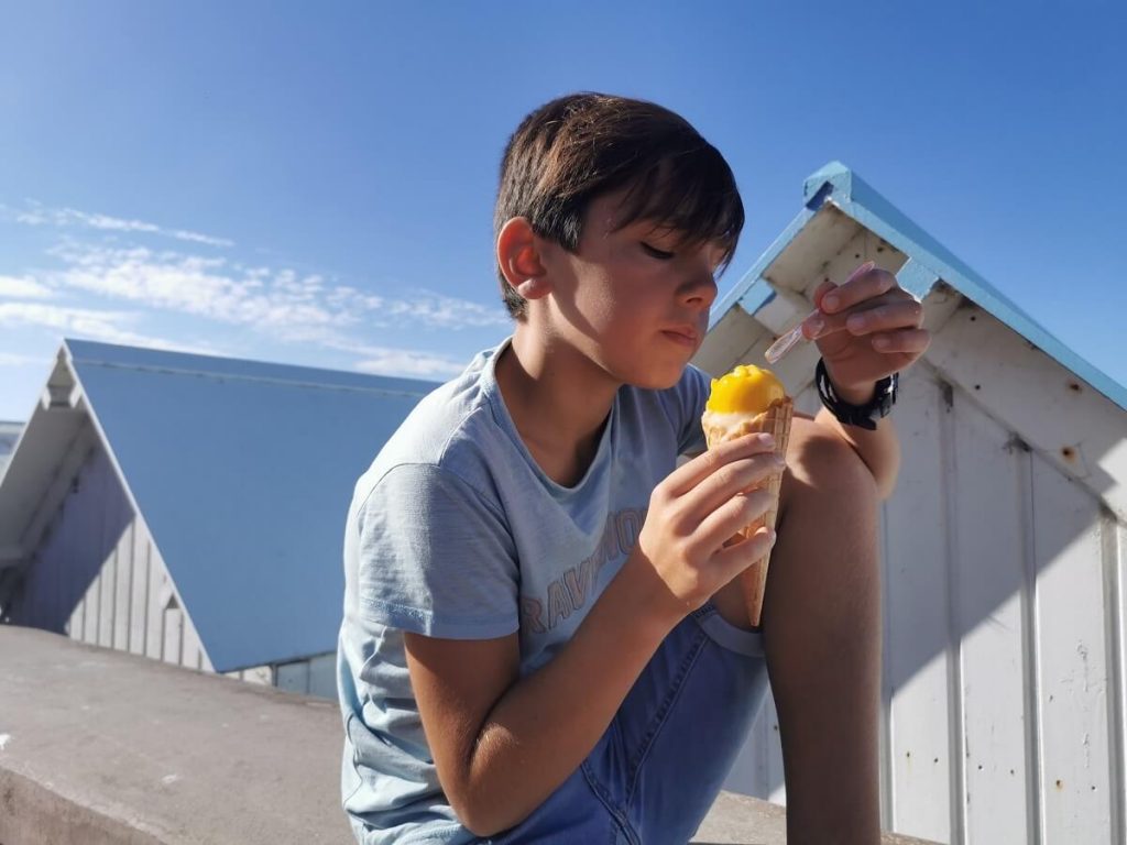 Assis sur le murée de la digue de Courseulles-sur-Mer, un jeune garçon déguste une glace couleur jaune soleil. Derrière lui, les toitures de deux cabines de plage posée sur le sable en contre-bas. Le ciel est bleu. – crédit : Nathalie Papouin