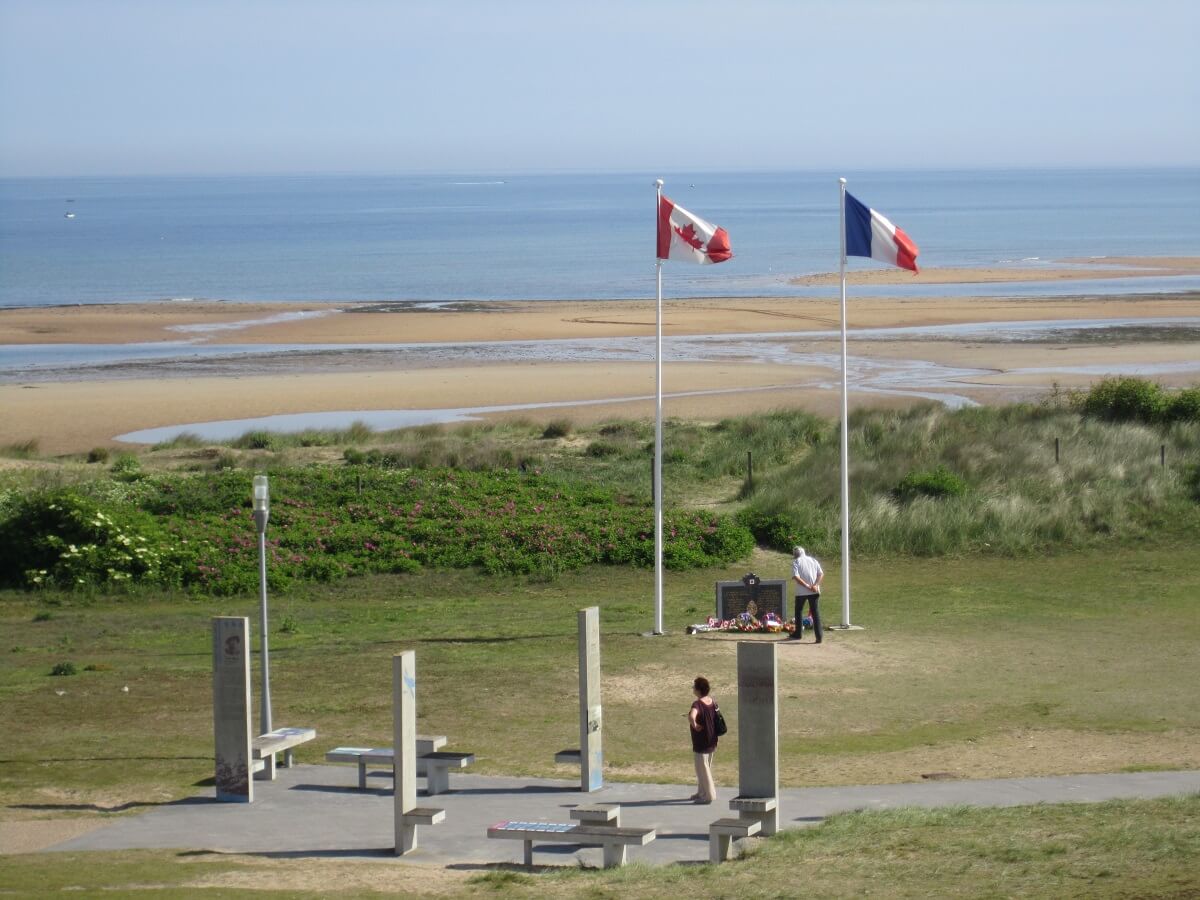 The photo was taken from a slightly lower angle, giving an overall view. In the foreground is part of Juno Park, located on the dunes, with the explanatory stelae and a picnic table. In the background, in the distance, between the French and Canadian flags, the stele of the Royal Canadian Navy. Finally, in the background, the Canadian beach stretches out, the sea is low.