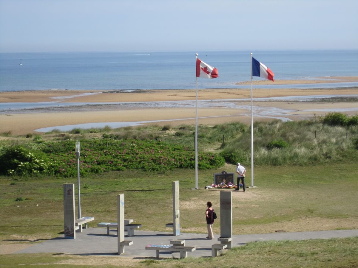 De foto is genomen vanuit een iets lagere hoek, waardoor je een totaalbeeld krijgt. Op de voorgrond is een deel van Juno Park, gelegen op de duinen, met de verklarende stèles en een picknicktafel. Op de achtergrond, in de verte, tussen de Franse en Canadese vlaggen, de stèle van de Royal Canadian Navy. Tot slot strekt zich op de achtergrond het Canadese strand uit, de zee staat laag.