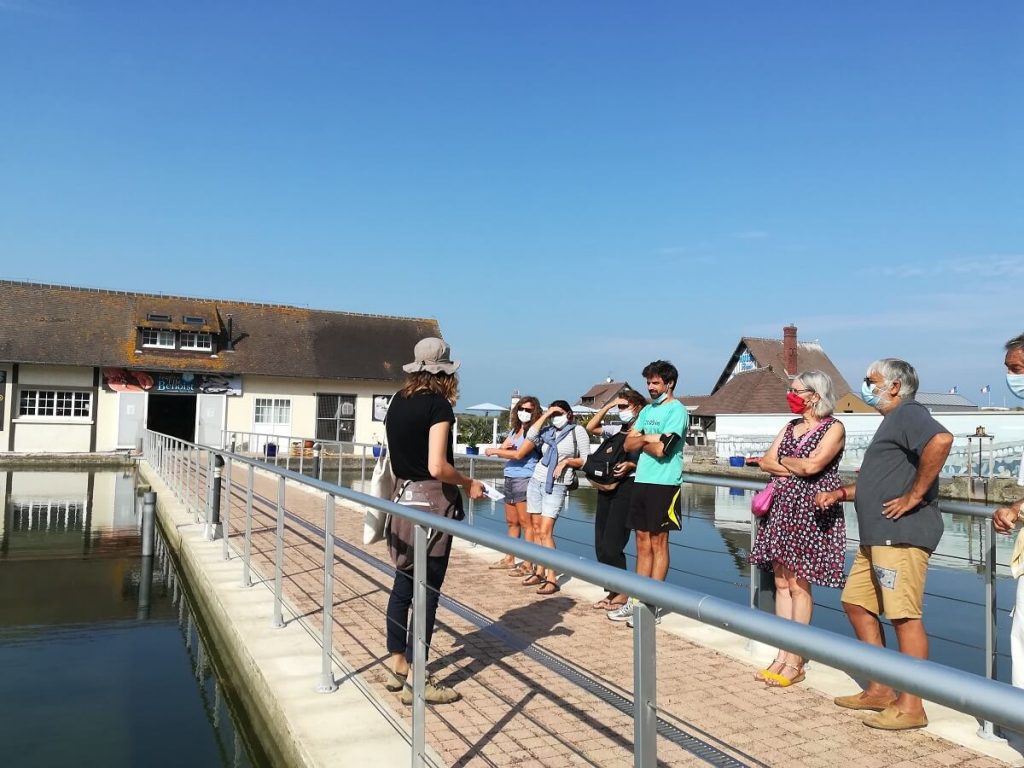 Adossé à la rambarde de l'allée qui traverse les parcs à huître de l'île de plaisance, un petit groupe de visiteurs écoute la visite guidée "Un port, une île, des huitres, une histoire courseullaise" animé par une des guides de l'Office de Tourisme.