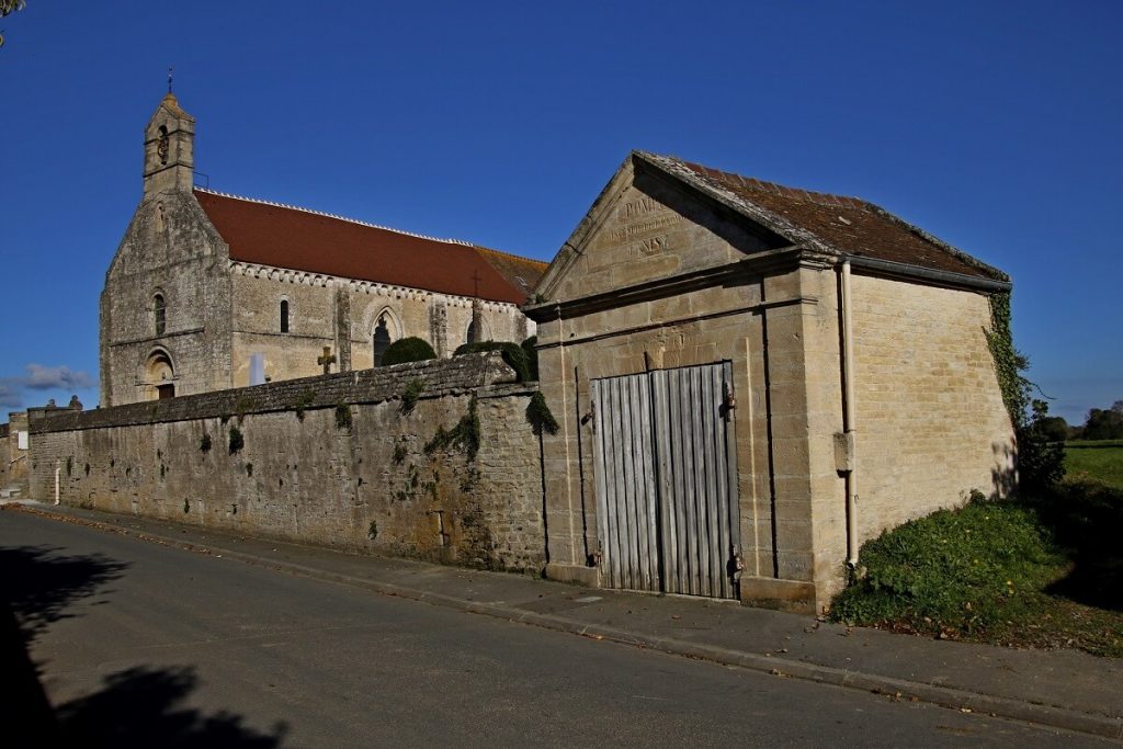 Prise depuis la rue, la photographie montre une partie de la façade latérale de l'église : une façade sobre percé de fenêtre à arcs brisés. Devant, on peut voir le mur du cimetière et dans son prolongement un petit bâtiment de style classique ornée d'une inscription sur son frontispice. - crédit Alain Lemarie