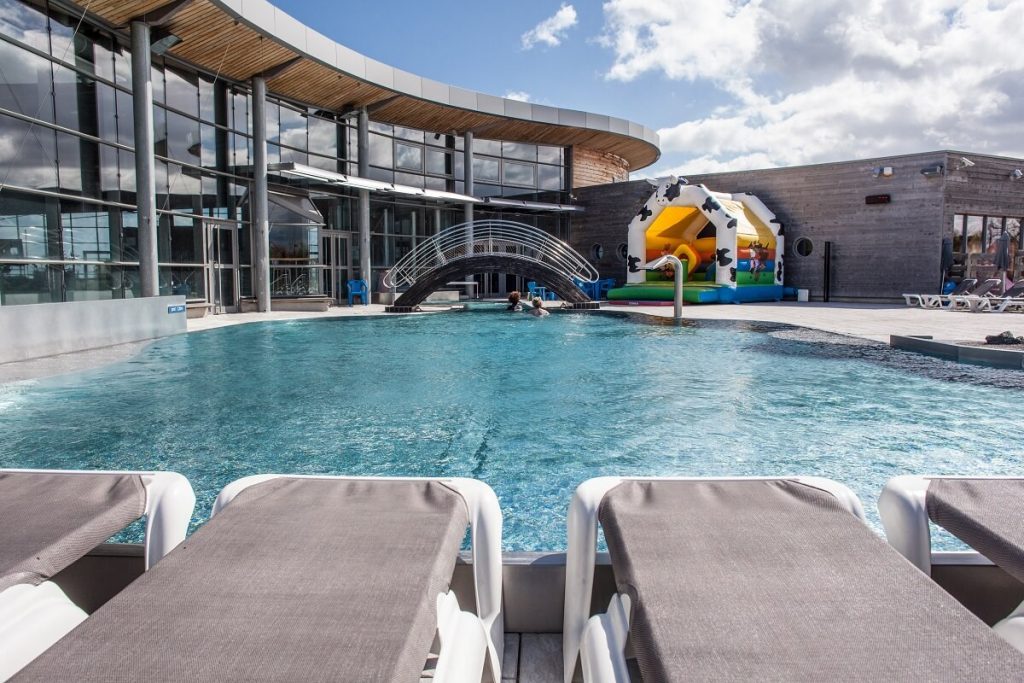 Het buitenzwembad van zwembad Aquanacre. Op de voorgrond de grijs-taupe ligstoelen voor het zwembad, op de achtergrond de houten brug die de ingang van het gebouw markeert en links de grote ramen van het zwembad. - credit: Aquanacre