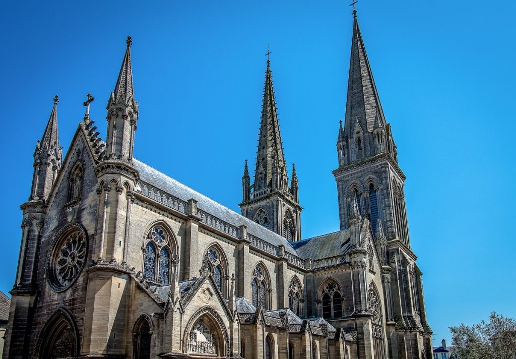La basilique Notre Dame de la Délivrande de style néo-gothique. La photo est prise de manière à voir le porche d'entrée, flanqué de deux tourelles, la façade latérale avec ses chapelles saillantes et les deux tours situées au niveau du chœur.