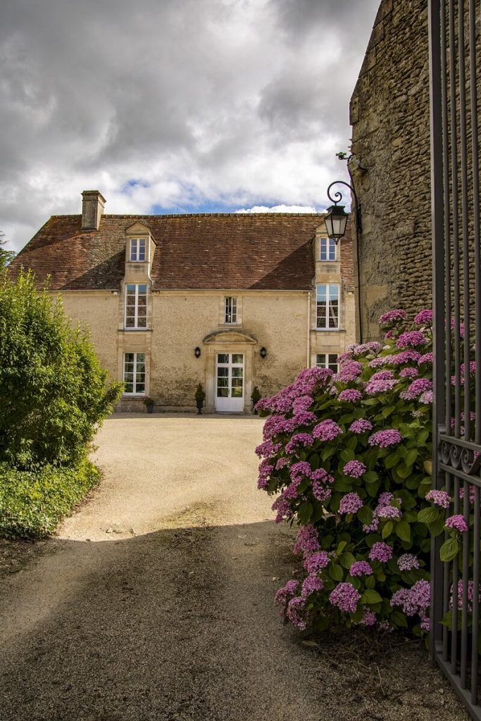 The photo shows a stone house in Creully. In the foreground is the entrance to the courtyard adorned with a flowering hydrangea, while in the background is a large house with a brown roof. The alignment of the 3-storey windows one above the other indicates a 19th or early 20th century building.