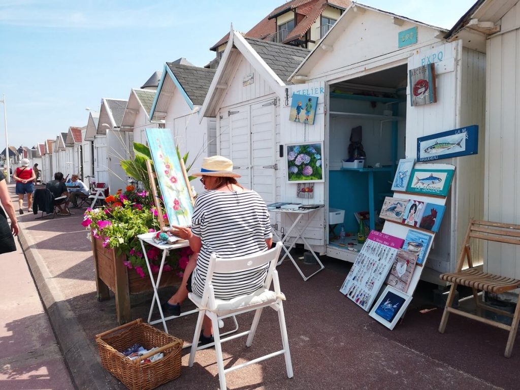 On the seafront, sitting in front of her beach hut, a painter paints a picture of flowers and exhibits her work.