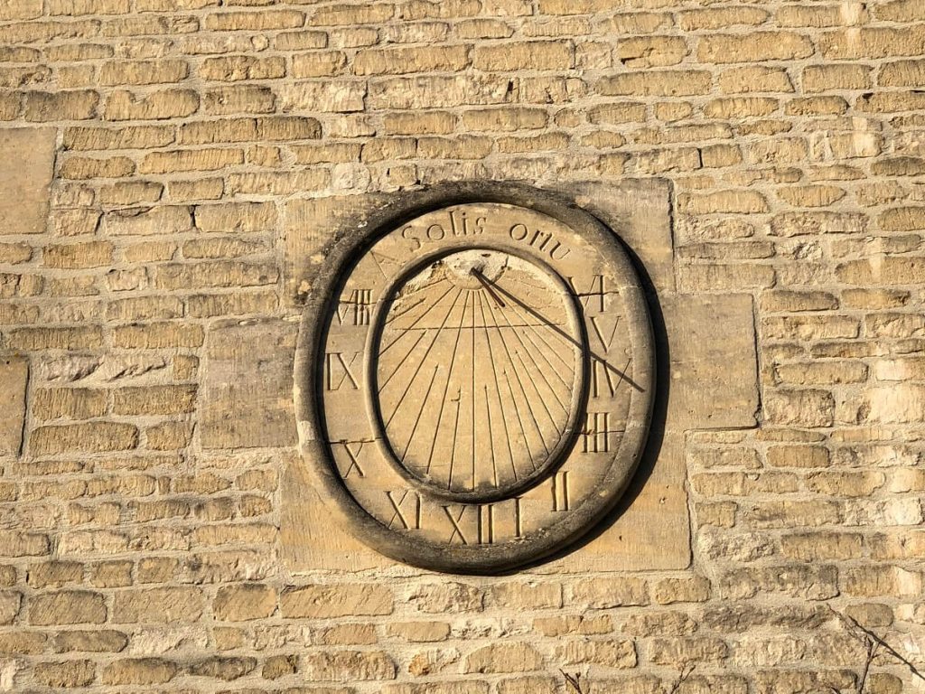 Op een okerkleurige stenen muur staat een zonnewijzer. Hij heeft de vorm van een medaillon: in het midden geeft de as waarvan de schaduw naar rechts valt 4 uur aan en daaromheen, omzoomd door twee uitstulpingen van steen, de uren in Romeinse cijfers. - credit: Mathilde Lelandais