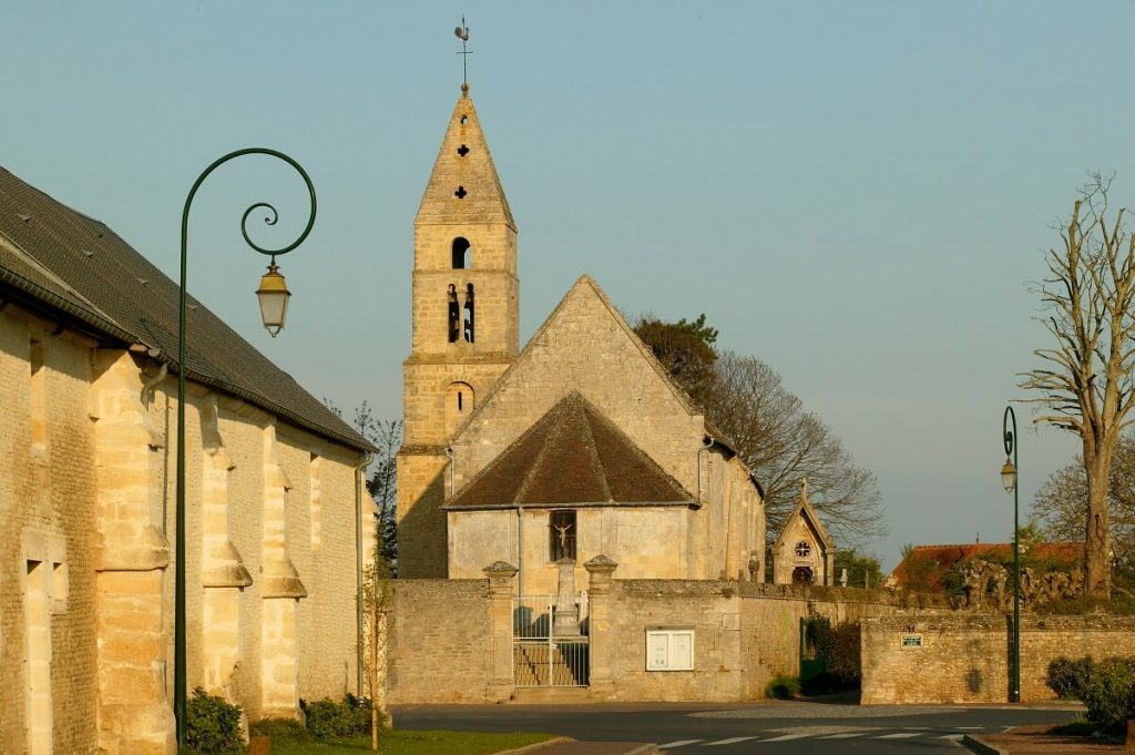 De kerk Saint-Martin in Colomby-Anguerny, genomen vanaf de straat, baadt in een zacht geel licht dat de okerkleur van de oude stenen goed doet uitkomen. Links is de stenen muur van een oud huis te zien en op de achtergrond, achter de kerkhofmuren, zie je de apsis en het schip van de kerk, evenals de klokkentoren, die iets uit het midden staat. De kerk heeft geen dwarsschip. - krediet Gregory Wait