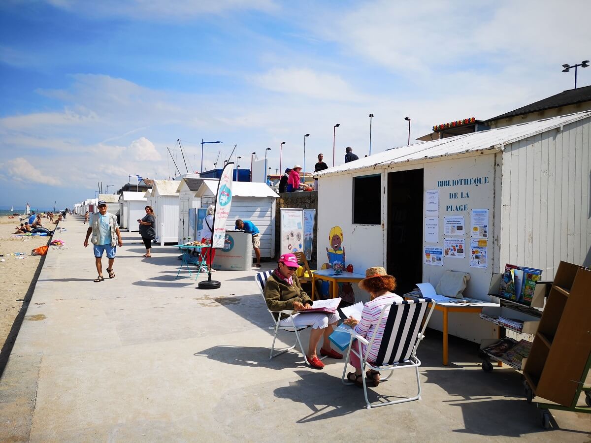 Under a blue sky sprinkled with white clouds, two readers sat in front of the "beach library" hut, with walkers on the sea wall and bathers on the beach in the distance.