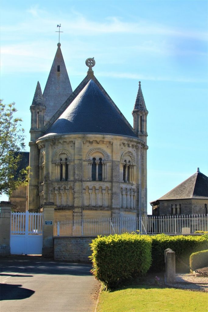 Au centre de la photo, prise depuis la rue, l'abside de l'église de Basly. Celle-ci est rythmée par des baies géminées plein cintres, deux tourelles encadrent l'abside surmonté d'un toit d'ardoise et d'une croix celtique. – crédit : Nathalie Papouin.