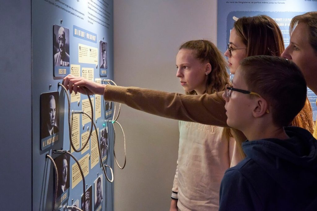The tour explores Juno dedicated to young people. On the left is the exhibition wall with photographs and texts to link together, on the right a young boy and two young girls look attentively at the panel, the taller of the girls has stretched out her arm to activate one of the interactive objects on the wall ©cjb ph.delval