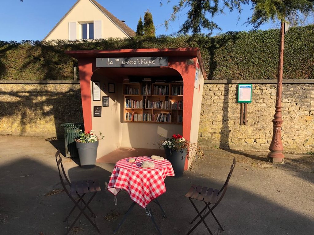 Geïnstalleerd in een oud betonnen bushokje, herschilderd in oranje en okergeel, de "Plumetothèque", een kleine zelfbedieningsbibliotheek, met op de voorgrond een kleine tuintafel met een rood en wit geruit tafelkleed en twee stoelen. - credit: Mathilde Lelandais