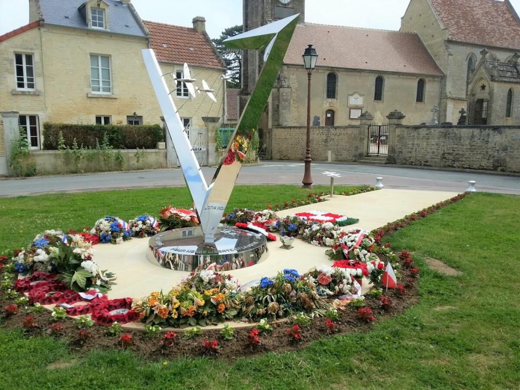 Le mémorial polonais de Plumetot entouré de gerbes de fleurs. Le ciel nuageux, la pelouse et les fleurs se reflètent dans le chrome de la sculpture en forme de "V". A l'arrière-plan des maisons en pierre et l'église. – crédit : A.M. Ruffier