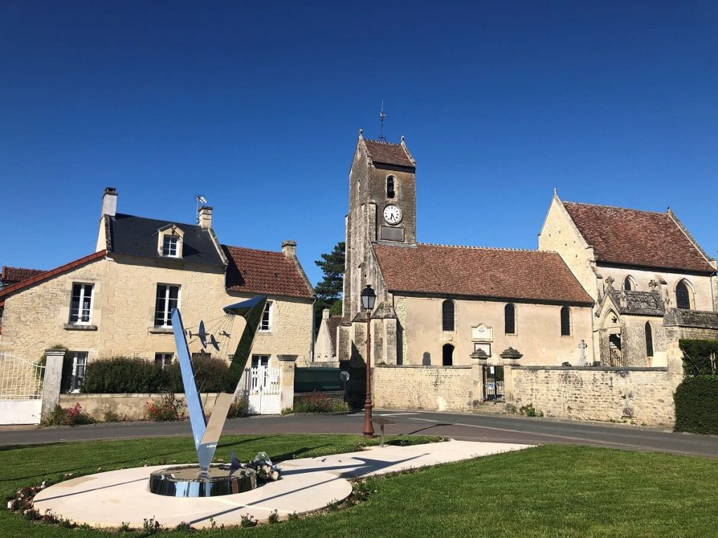 Au premier plan sur la gauche la sculpture du mémorial polonais de Plumetot : elle a la forme d'un "V" majuscule dont le haut de la hampe droite est recourbé vers l'intérieur, formant un angle saillant. La sculpture est chromée et reflète le paysage comme un miroir. A l'arrière-plan des maisons en pierre et l'église de Plumetot. – crédit : Mathilde Lelandais