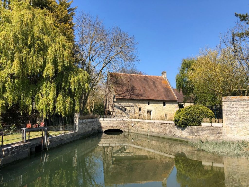 La photo est prise depuis le pont de l'allée Bouchon : au premier plan la Seulles bordée à cet endroit de murs de pierre, à l'arrière-plan, au centre de l'image, le moulin de Courseulles qui se présente sous la forme typique d'une maison en pierre à la toiture de tuiles. Sur la gauche, au deuxième plan un saule pleureur et une écluse. – crédit : Mathilde Lelandais