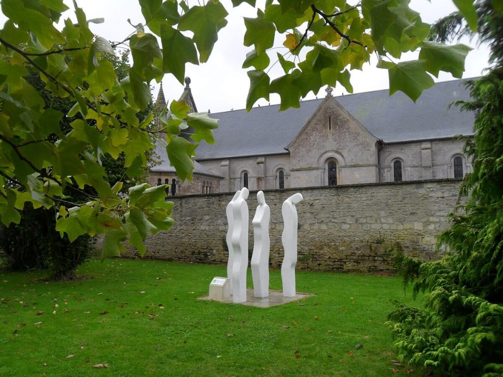 In the background, the side facade of Basly church, partly hidden by the cemetery wall. In front of this wall, set in the middle of a green lawn, is a sculpture by Serge Saint, consisting of three white silhouettes, with only the curves formed by the backs and faces in relief (the sides of the silhouettes are flat). You can make out 3 men, two of whom are looking at a woman, and the third, standing apart, who has his back to the group.