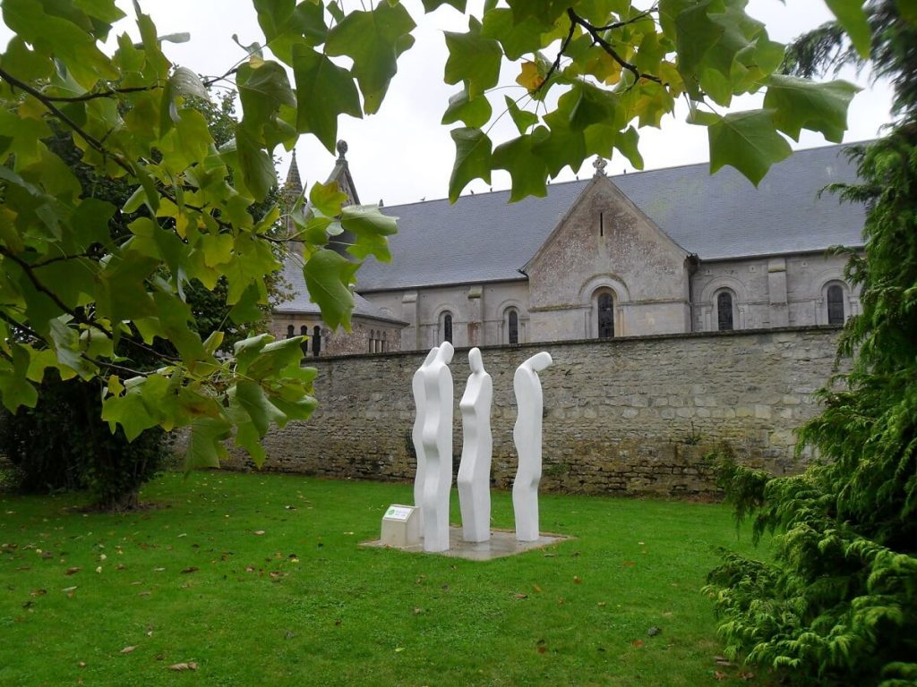 A l'arrière-plan la façade latérale de l'église de Basly, en partie cachée par le mur du cimetière. Devant ce mur, posée au milieu d'une pelouse verdoyante une sculpture de Serge Saint, constitué de trois silhouettes blanches, seules les courbes formées par les dos et les faces sont mises en relief (le côté des silhouettes est plat). On peut distinguer 3 hommes, deux qui regardent une femme, et le troisième, à l'écart, qui tourne le dos au groupe.