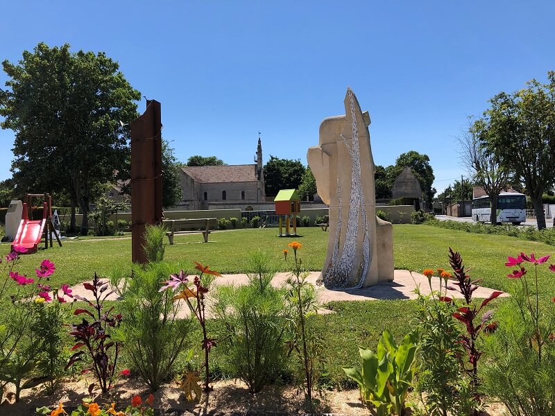 In the foreground, a contemporary work of art made up of two sculptures: one in metal, the other in stone and mosaic, symbolising the passage from oppression to freedom. In the background, a slide and a book hut, with the church of Cresserons in the distance.