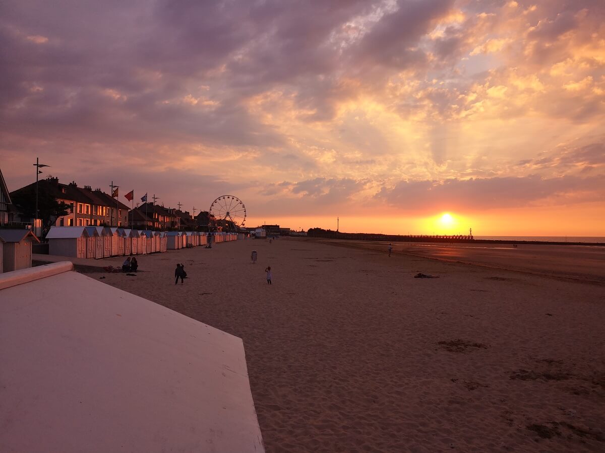 De avond valt op het strand van Courseulles-sur-Mer. In de verte, rechts op de foto, baadt een gele zon boven de steiger de lucht, onderbroken door grijs-mauve wolken, in haar oranje stralen. Links weerspiegelen de hutten en ramen van de huizen op de zeedijk de oranje gloed van de zonsondergang. Links op de achtergrond zijn de spaken van het reuzenrad te zien, terwijl het fijne zandstrand zich over de rest van de foto uitstrekt, met een paar wandelaars die er nog rondhangen.