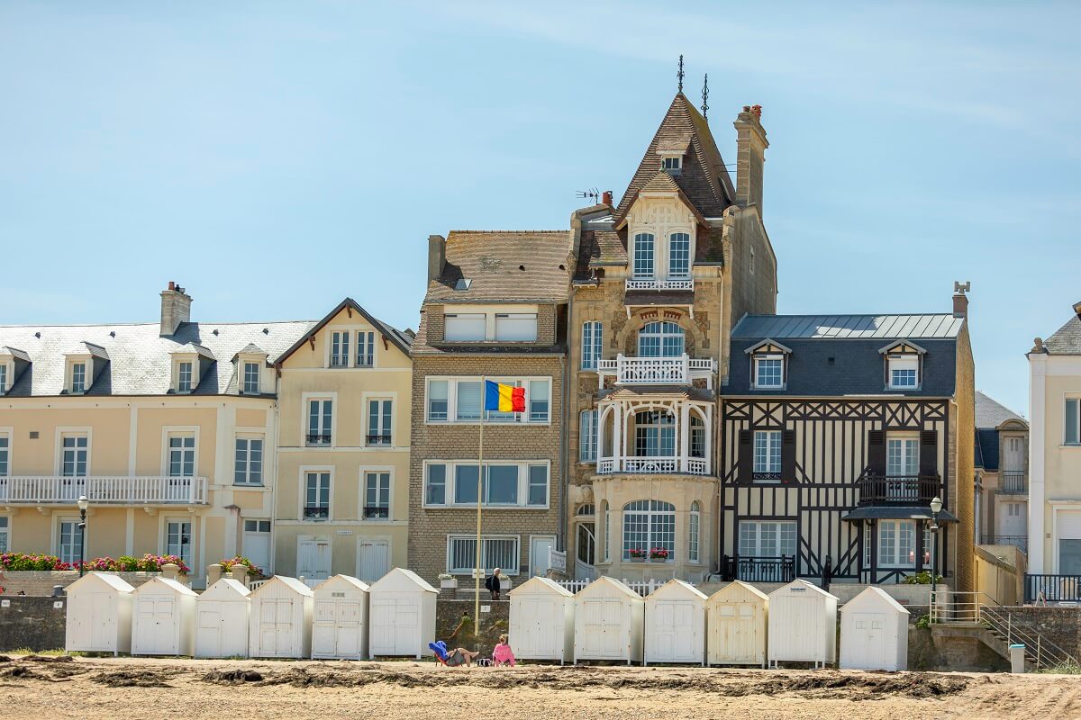 Sous un ciel bleu, la plage de Saint-Aubin-sur-Mer avec ses cabines blanches allignées contre le mur de la digue, et à l'arrière plan une des villas belle époque de la ville, la Loggia, à la façade étroite composée d'un balcon  et d'une baie arqué (bow-window).