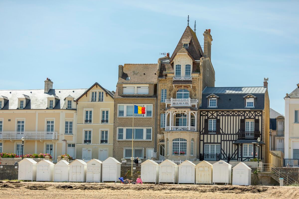 Onder een blauwe hemel, het strand van Saint-Aubin-sur-Mer met zijn witte hutten tegen de muur van de zeewering, en op de achtergrond een van de villa's uit de belle époque van de stad, de Loggia, met zijn smalle gevel die bestaat uit een balkon en een boograam.