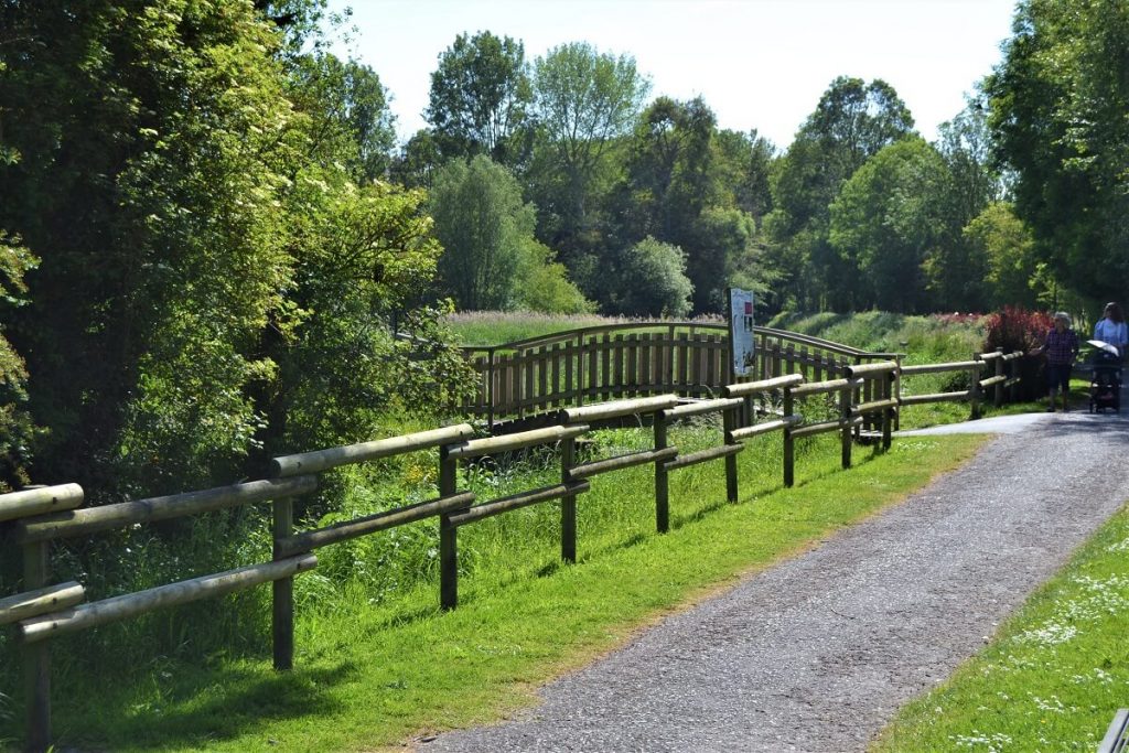 La promenade Thomas : un petit chemin de graviers entouré d'herbe et de bosquets, balisée par une barrière en bois. A l'arrière-plan, sur la gauche un petit pont de bois passe au-dessus de la Douvette.