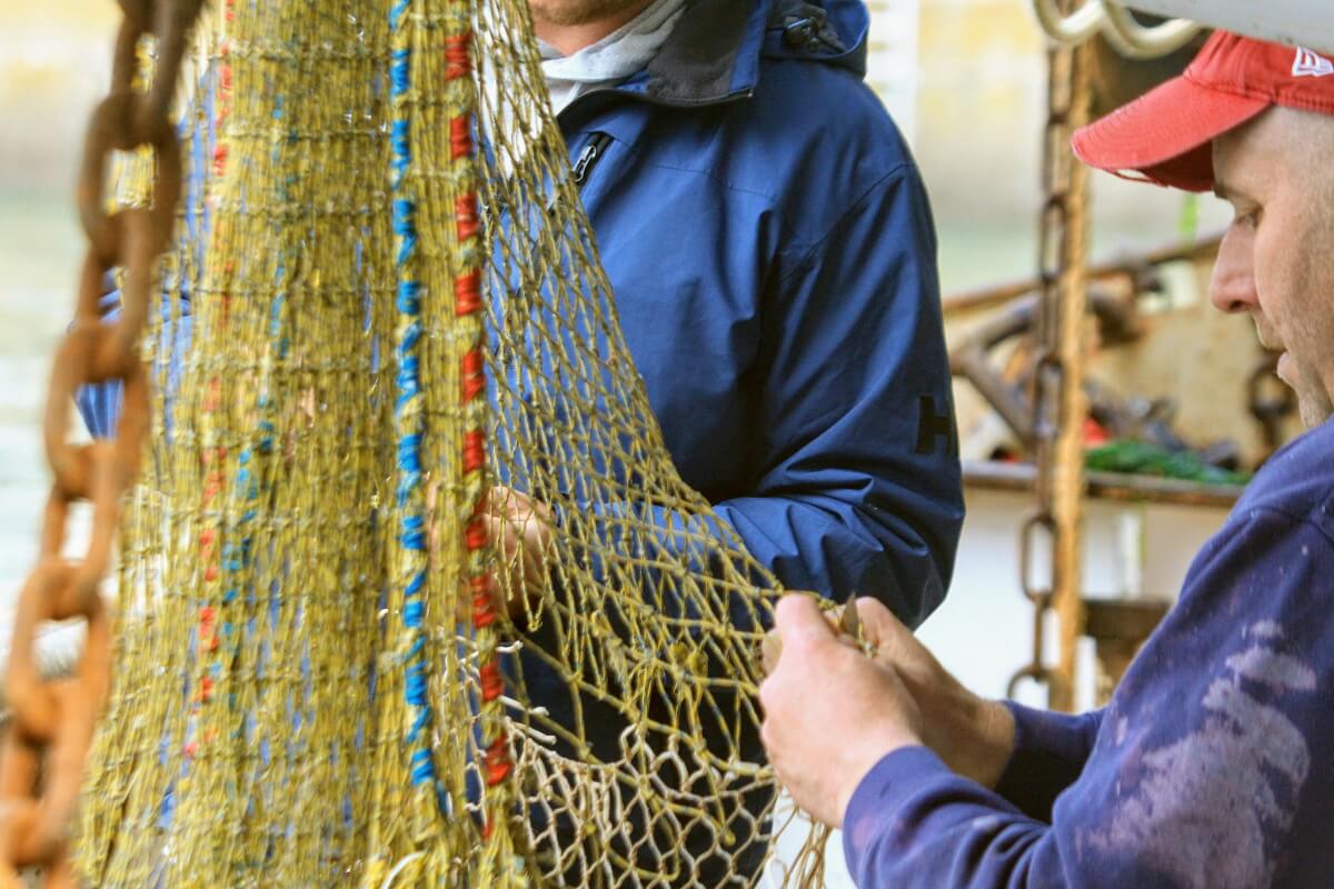 Two people mending a fishing net. On the left of the picture is the yellow net, the edges of which are reinforced with red or blue cords, while on the right the hands are busy repairing the meshes.