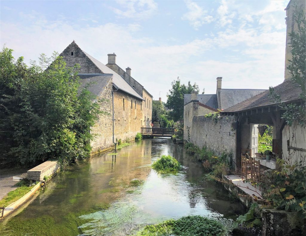 De rivier de Mue gezien vanaf de kleine brug bij Place du Planître, met Creully stenen huizen langs de rivier.