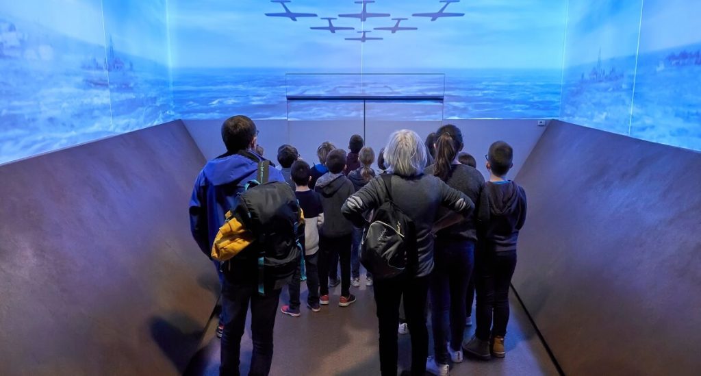 The entrance to the Juno Centre museum. The room is laid out in such a way as to give visitors the impression of being on one of the barges at D-Day. Here, a group of visitors watch the film projected on the walls while waiting for the museum doors to open. ©cjb ph.delval