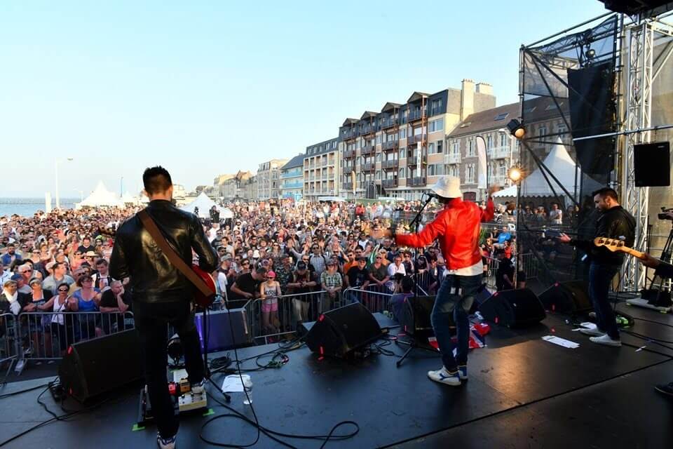 De foto is genomen vanaf het Tribute Fest podium in 2019 en toont de gitaristen en de zanger, in het midden en met rode jassen aan, in volle uitvoering voor het publiek dat naar het concert kwam kijken. - krediet: Didier Mignon