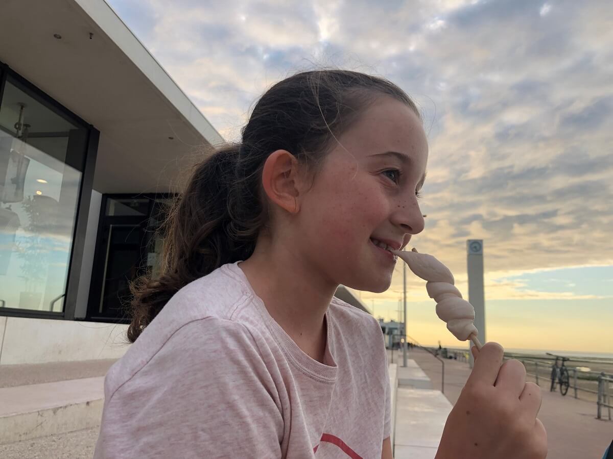 A young girl takes a bite out of a guigui, as evening falls and the great clock on the Luc-sur-Mer seawall can be seen in the distance behind her. - credit: Mathilde Lelandais