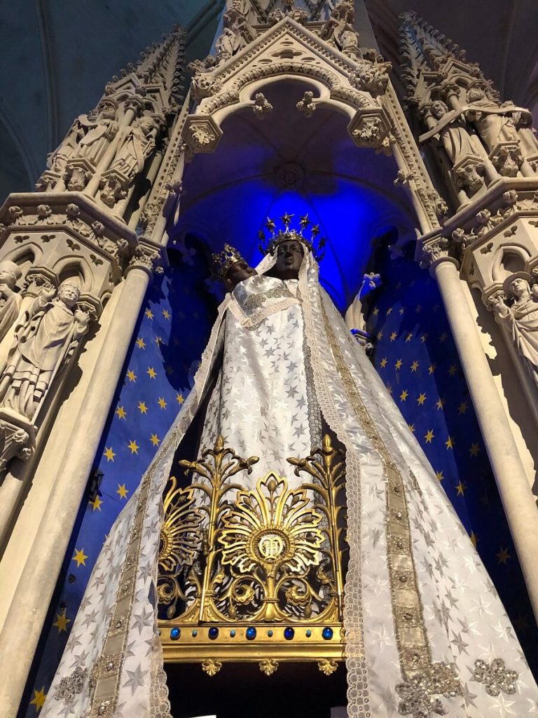 La vierge noire de la basilique de Douvres-la-Délivrande. La statue est prise en contre plongée, habillée d'une longue robe et d'un voile somptuaire blanc brodé d'étoiles argenté. Elle est posée sur une couronne d'or orné de pierres bleu, dans une niche sculptée en style néo-gothique flamboyant. Le fond de la niche est tapissé d'un tissu bleu nuit étoilé. - crédit : Mathilde Lelandais