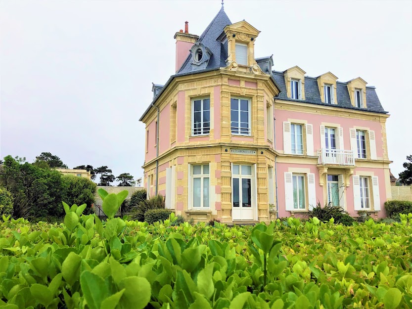 Villa Les Hirondelles, Saint-Aubin-sur-Mer. In the foreground, a well-trimmed light-green hedge gives the impression of a blanket of greenery, from which emerges the pink-rendered villa, composed of two wings: the one facing the camera features an arched façade. The window surrounds are in yellow ochre stone, the roof in slate. - credit: Mathilde Lelandais