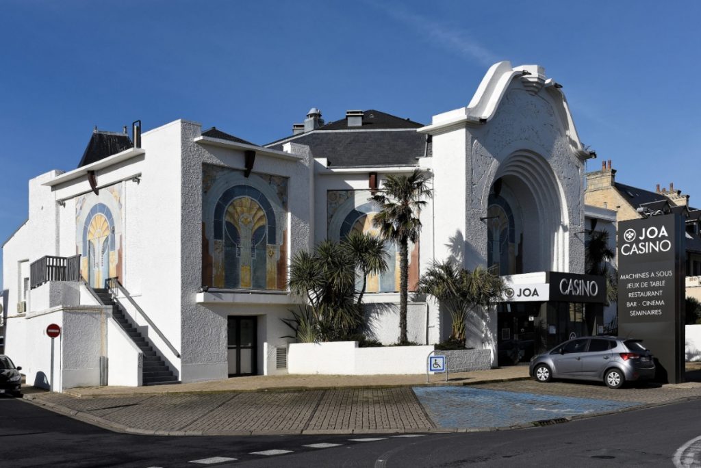 The Joa casino: in art-deco style, the photograph shows the side facades, decorated with a large mosaic motif in blue, golden ochre and yellow, red ochre. The building's walls are white, and the entrance, on the right of the photo, takes the form of a huge porch (several storeys high). - credit: François Dupont