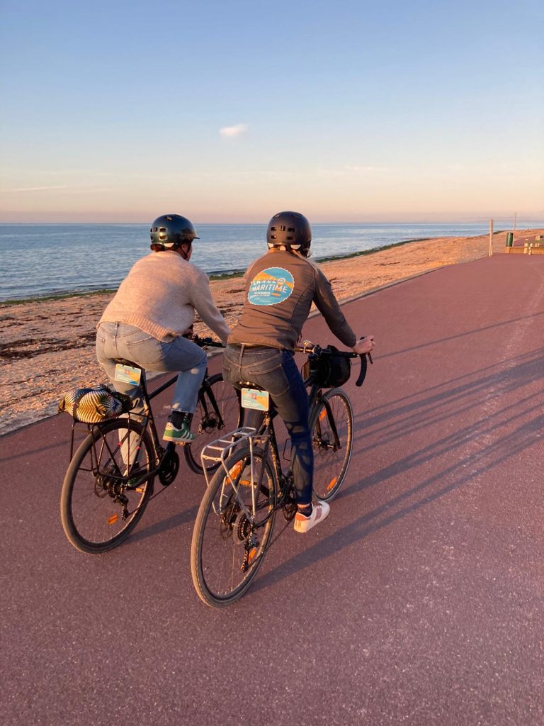 Deux cyclistes en balade sur la vélomaritime à Courseulles. On les voit de dos, côte à côte, directement sur leur gauche la plage et la mer. Le soleil de la fin d'après-midi étant leurs ombres devant eux, la mer et haute et bleu, le ciel aussi. - crédit : les valises de Sarah, Calvados attractivité.