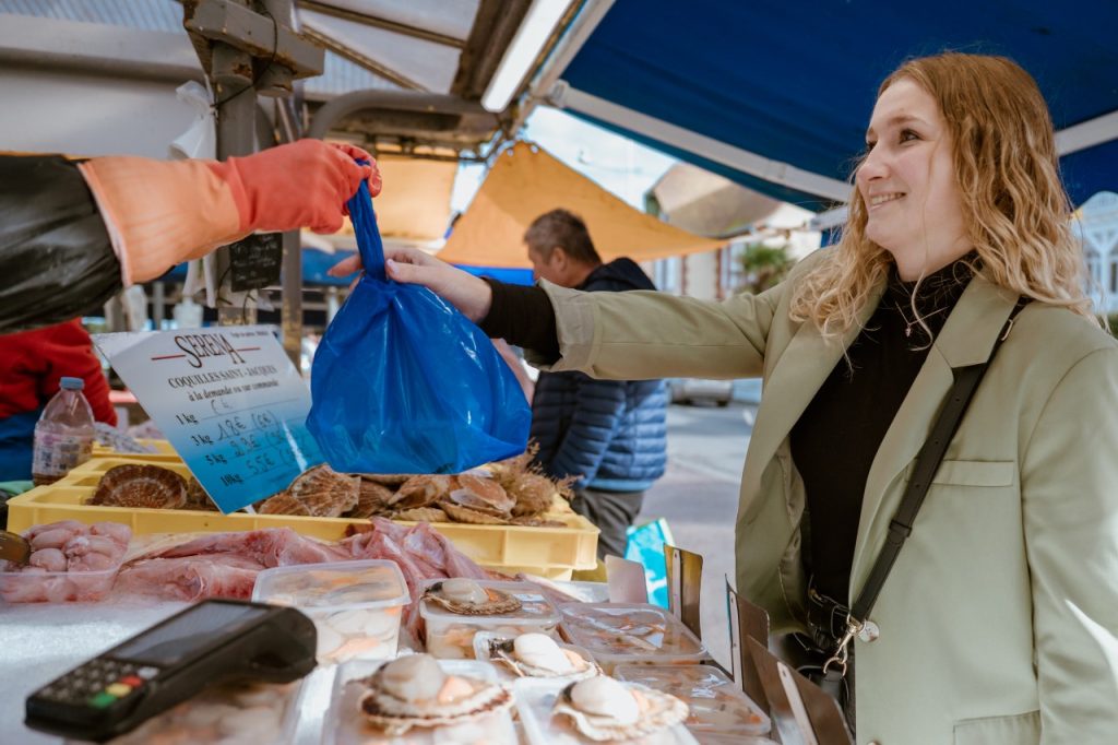 Une cliente souriante reçoit un sac bleu de produits frais à un étal du marché aux poissons de Courseulles-sur-Mer, avec des coquilles Saint-Jacques au premier plan.