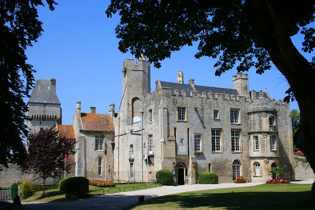 Façade en pierre du Château de Creully avec sa tour carrée, ses fenêtres à meneaux et ses jardins fleuris.
