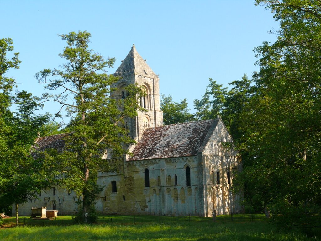Église romane de Thaon au milieu des arbres avec son clocher à pyramide en pierre calcaire sous un ciel bleu.