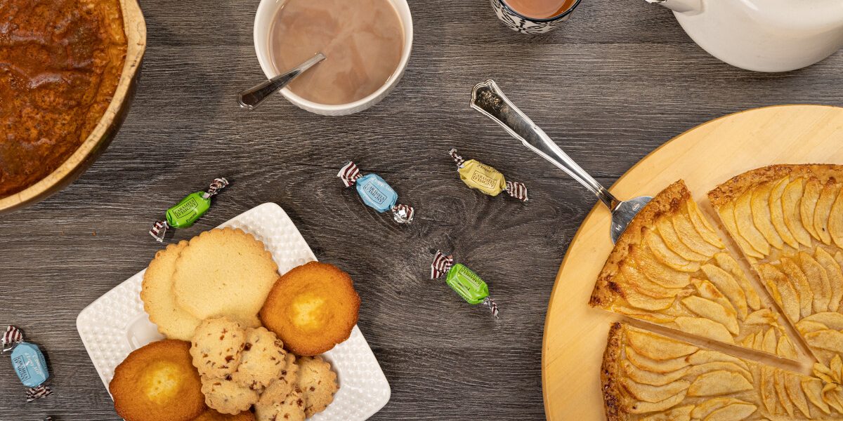Vue de dessus (flat lay) d'une table en bois sombre garnie d'un goûter normand : une tarte aux pommes tranchée sur un plateau circulaire, une assiette de madeleines et de sablés, des caramels d'Isigny éparpillés, un bol de chocolat chaud, une tasse de thé et une théière blanche.