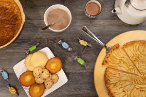 Vue de dessus (flat lay) d'une table en bois sombre garnie d'un goûter normand : une tarte aux pommes tranchée sur un plateau circulaire, une assiette de madeleines et de sablés, des caramels d'Isigny éparpillés, un bol de chocolat chaud, une tasse de thé et une théière blanche.
