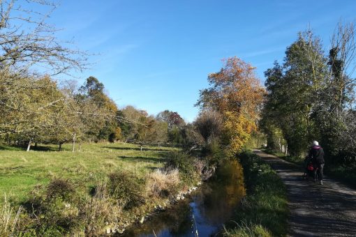 Une petite rivière calme bordée d'arbres aux couleurs d'automne, avec un chemin de terre à droite où marche une personne.