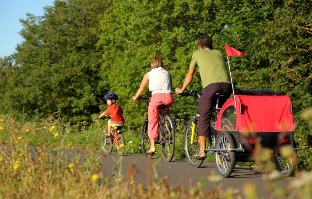 Parents à vélo avec une remorque rouge pour enfant sur une voie goudronnée en pleine nature.