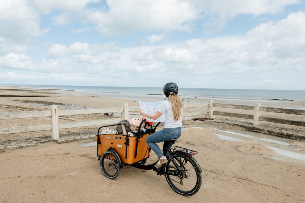 Femme à vélo avec deux enfants dans un vélo-cargo sur la digue de Luc-sur-Mer face à la mer.